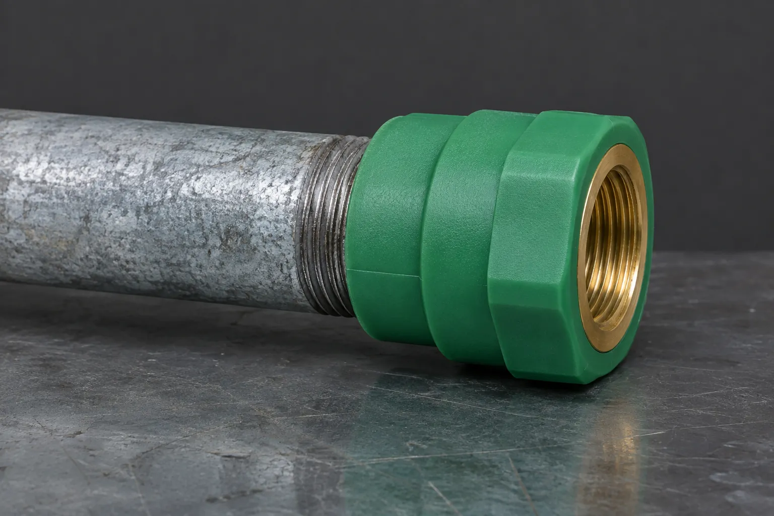 Green PP-R fitting with a brass female threaded insert screwed onto the freshly cut and threaded end of a grey galvanised steel water pipe on a steel workbench