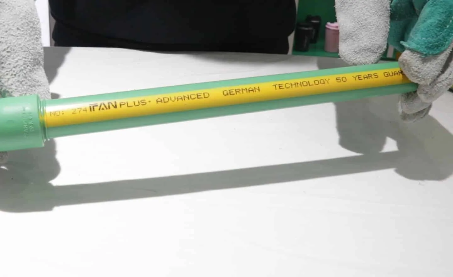 Gloved hands holding a green PP-R pipe with a yellow stripe printed NO: 274 IFAN PLUS ADVANCED GERMAN TECHNOLOGY 50 YEARS